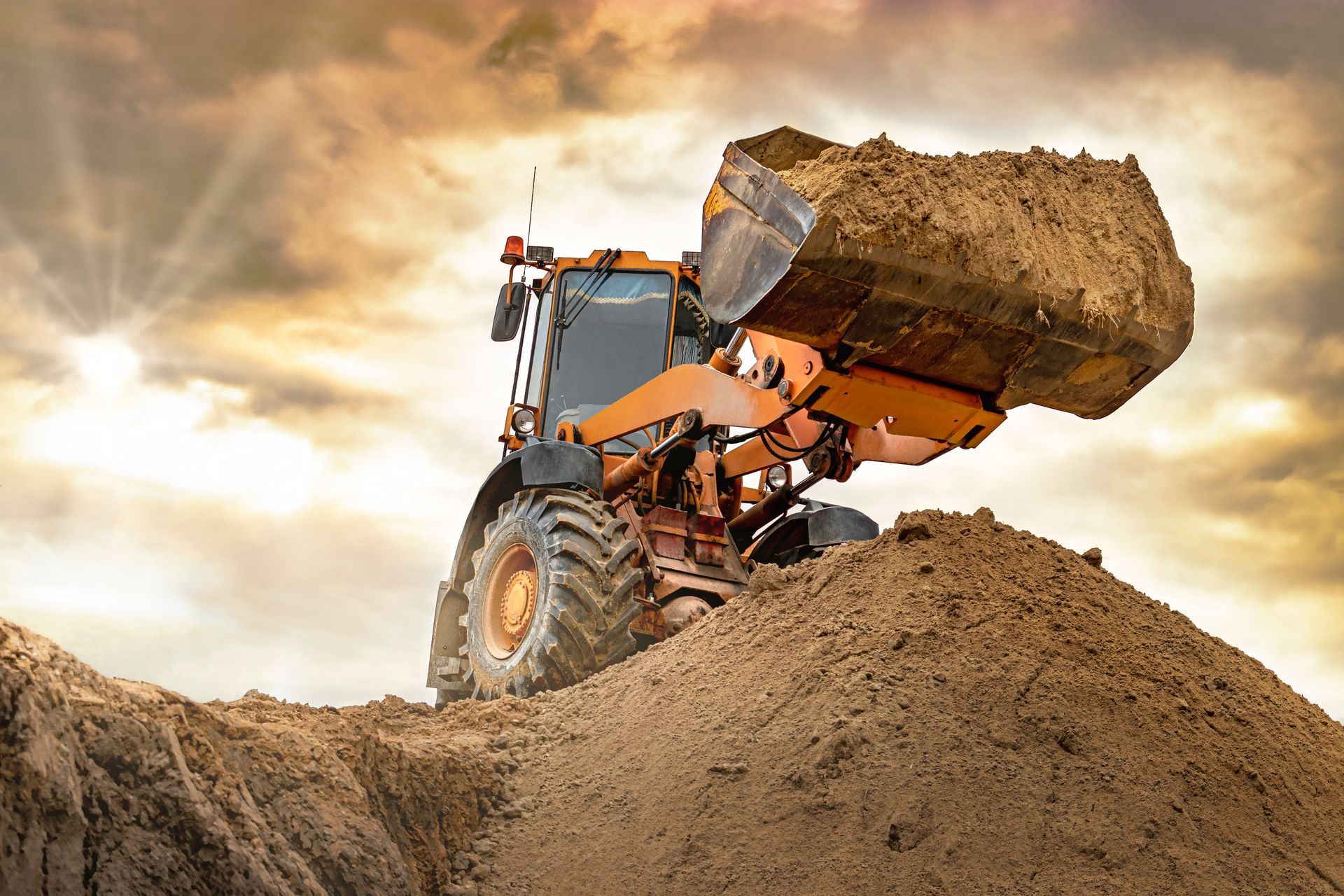 Orange backhoe loading a mound of dirt against a cloudy sky.