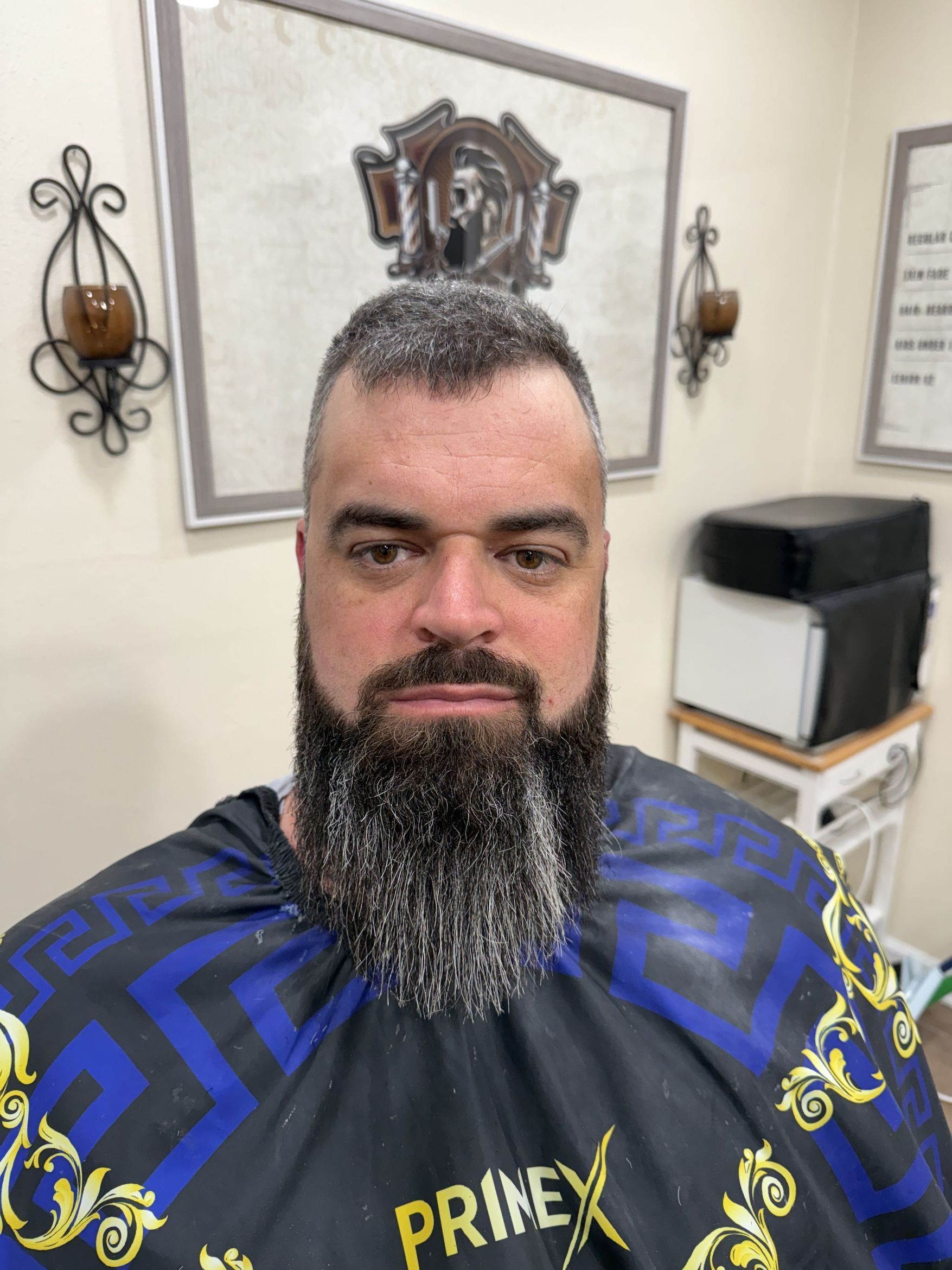 A person with a thick, graying beard wears a dark, patterned barber cape in a salon with wall art.