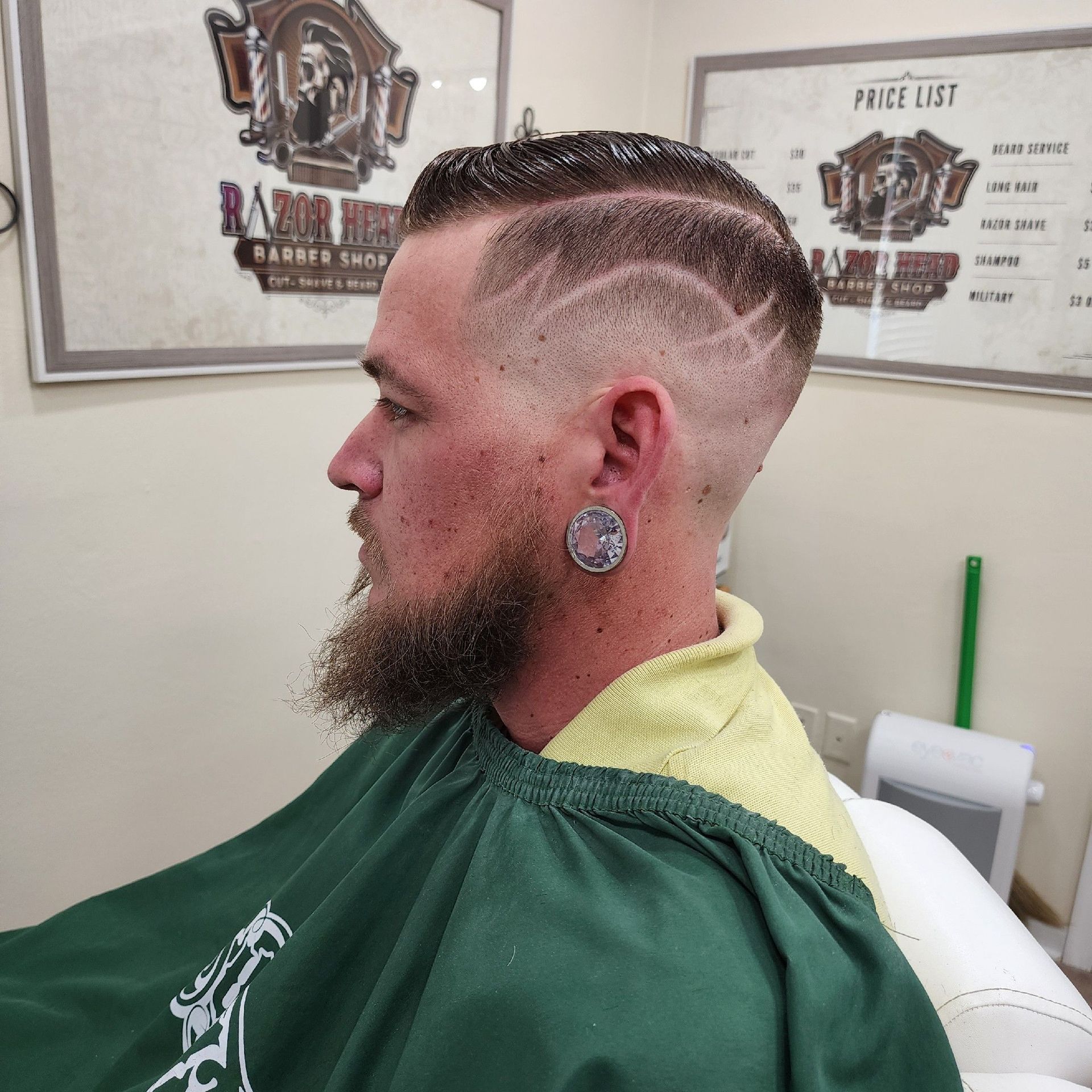 Side profile of a person with a skin fade haircut featuring a carved wave design, beard, and ear gauge in a barbershop.