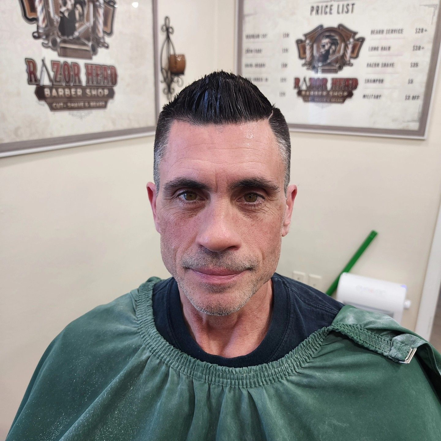A man with a short, spiky haircut and green barber cape, centered in front of a shop's logo and price list.