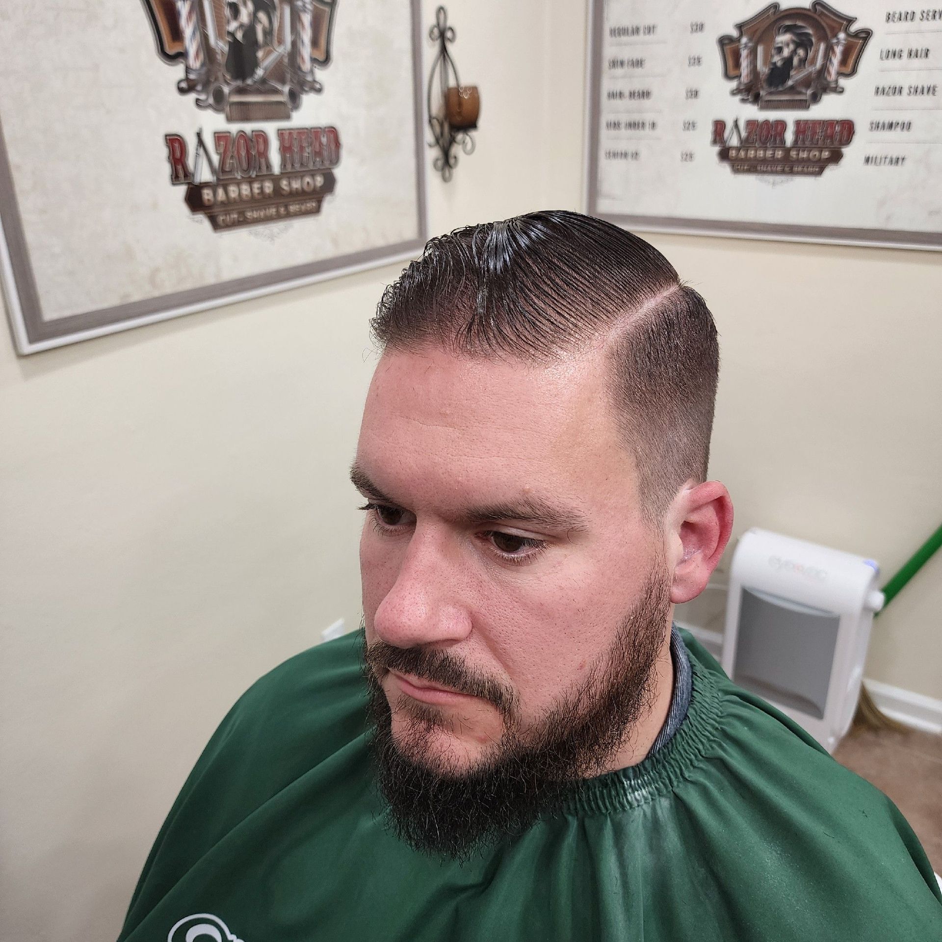 A person with a short, clean-cut fade hairstyle and a groomed beard sits in a barbershop wearing a green cape.