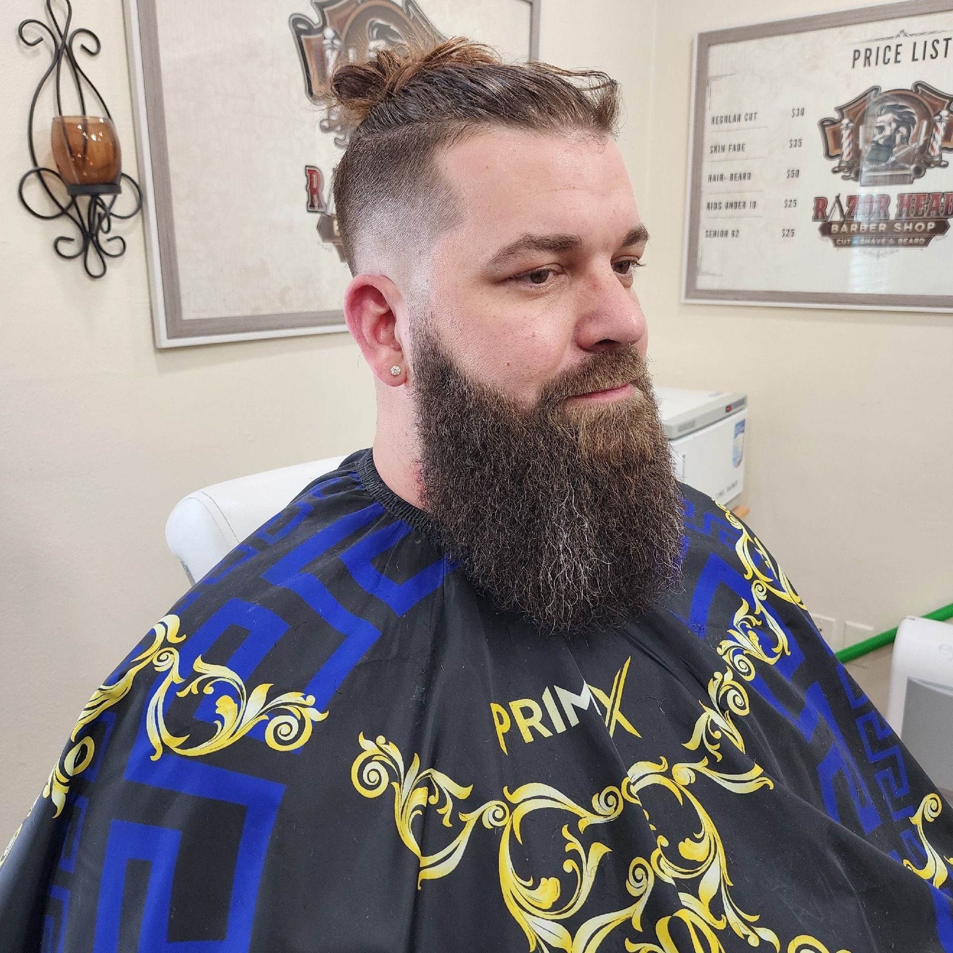A man with a groomed beard and a top-knot haircut sits in a barber cape at a barbershop.