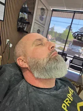 Bearded man reclining in a salon chair with eyes closed, in a barbershop with mirrors and window view behind him.