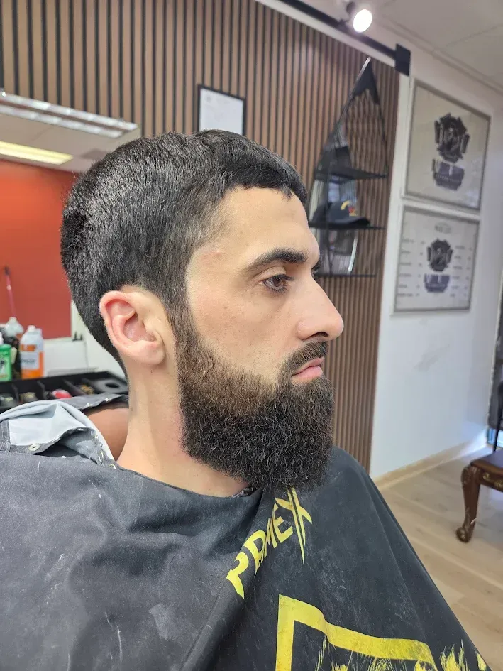 Man with dark beard in a barbershop chair, looking left during a haircut.