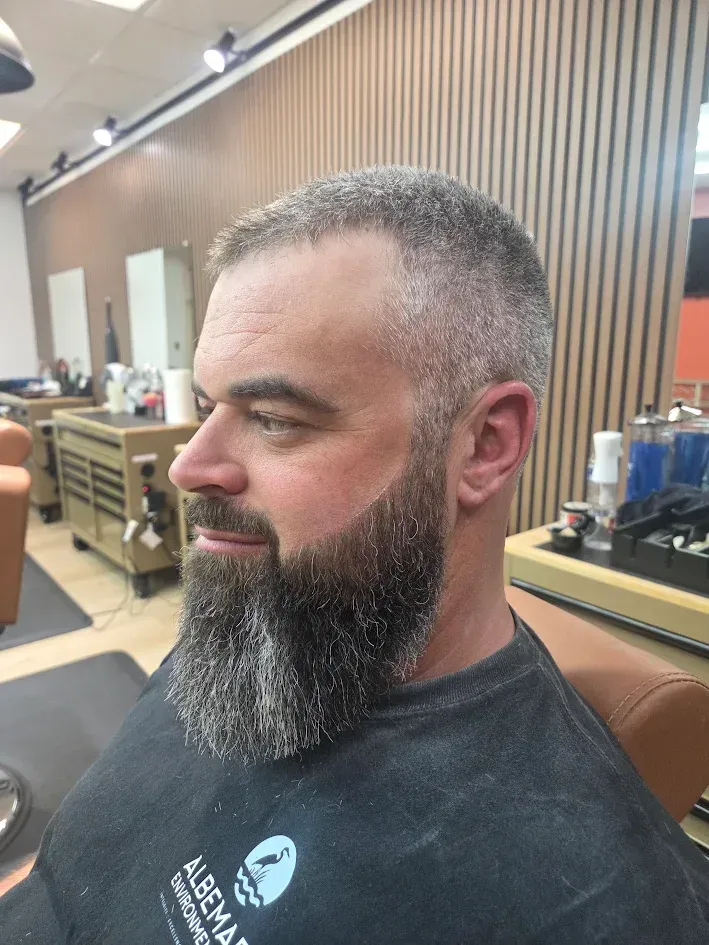 Man with a full beard sitting in a barber chair in a barbershop, wearing a black cape.