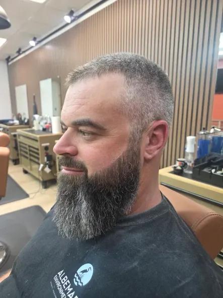 A barber in a white shirt and black apron trims a client’s mustache with scissors in a professional barbershop.