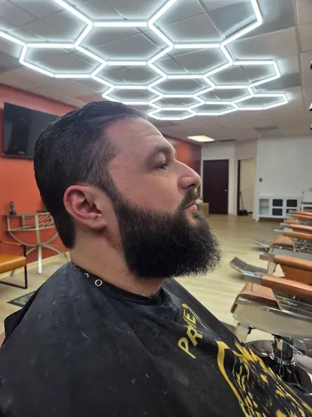 A barber in a white shirt and leather apron uses a straight razor to style a geometric design into a client's hair.