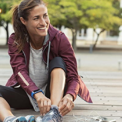 Woman tying running shoe while sitting on a wooden path in a park, smiling in a maroon jacket.