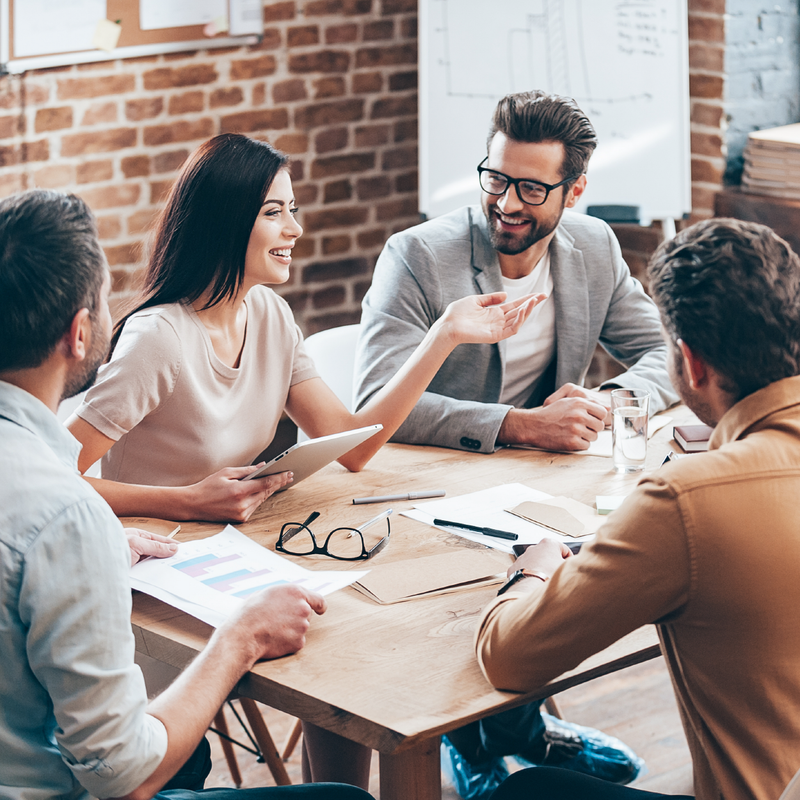 Team meeting in a bright office, with four people talking around a wooden table covered in papers and laptops.
