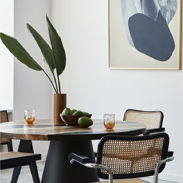 Modern dining room with round wooden table, woven chairs, vase of leaves, and abstract wall art
