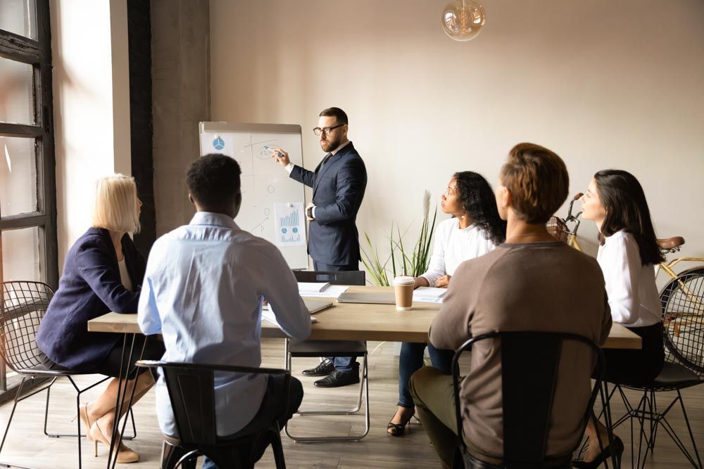Business presentation in a meeting room, with a presenter speaking to seated colleagues by a whiteboard.