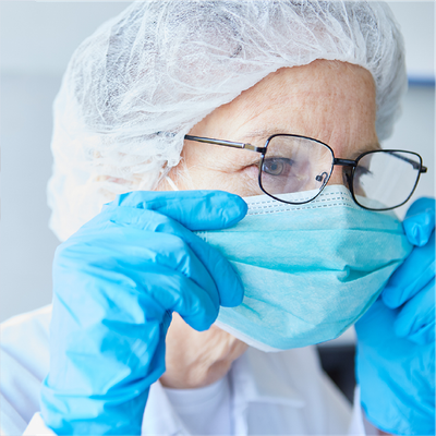 Healthcare worker adjusting a light blue surgical mask and glasses, wearing blue gloves and a hairnet