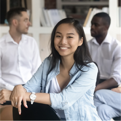 Smiling woman in a light blue shirt sitting indoors with two people blurred in the background