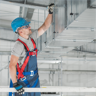 Worker in blue hard hat and safety harness inspecting ceiling ductwork on a construction site
