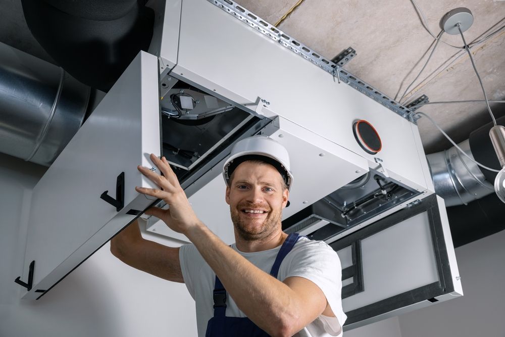 Technician in a white hard hat smiles while opening an overhead HVAC ceiling panel in a utility room
