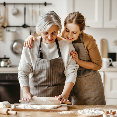 Two women in aprons roll dough together in a bright kitchen, smiling beside a floured counter.