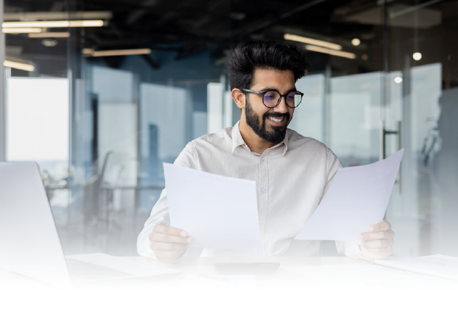 Smiling man in glasses reviewing papers at a bright office desk