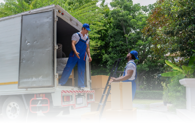 Two workers in blue uniforms unload a white truck using a hand truck outdoors.