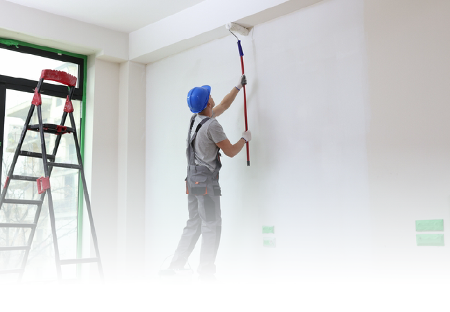Painter on a ladder rolling white paint onto a wall in a bright room