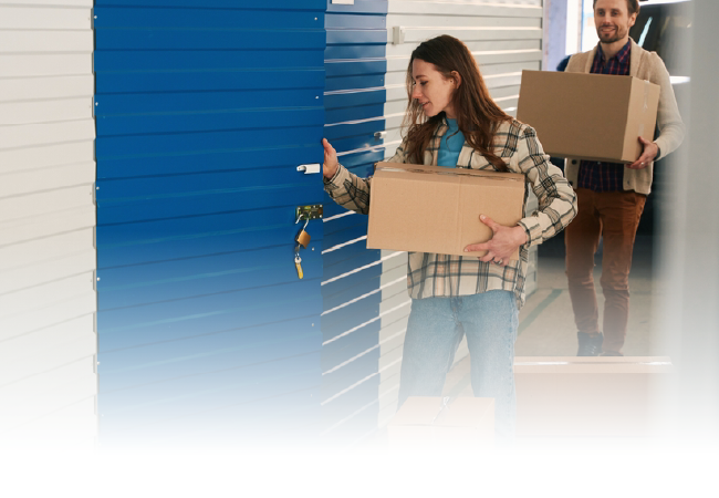 People carrying boxes near a blue storage unit door, moving into or out of a building.
