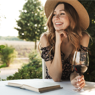 Smiling woman in a straw hat with a glass of red wine, seated by an open notebook outdoors