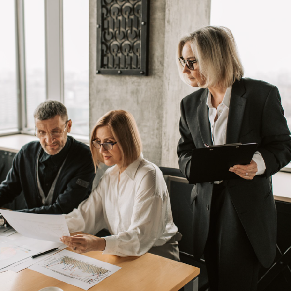Three coworkers review documents at a desk while a woman in a black suit holds a clipboard nearby