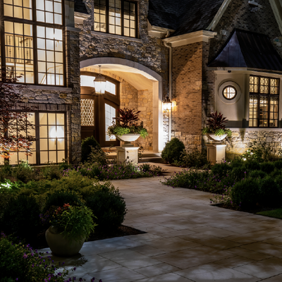 Illuminated stone house courtyard with arched entryway, tall windows, and potted plants at night