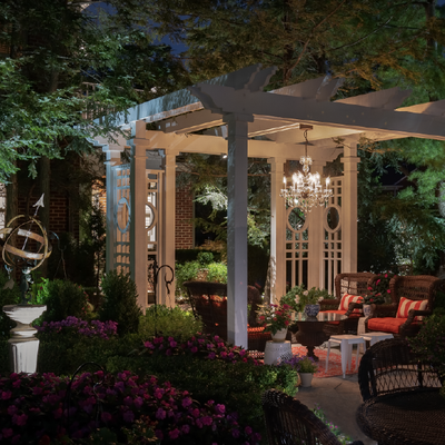 Cozy garden patio at dusk with white pergola, chandeliers, and red cushioned seating amid flowers and trees