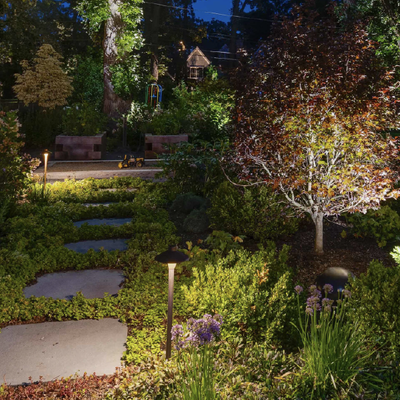 Lit garden path with stepping stones, glowing lamp posts, and a small tree beside lush greenery at night