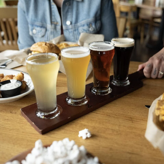 Four craft beers on a wooden tray at a bar table, with food and a person seated nearby.