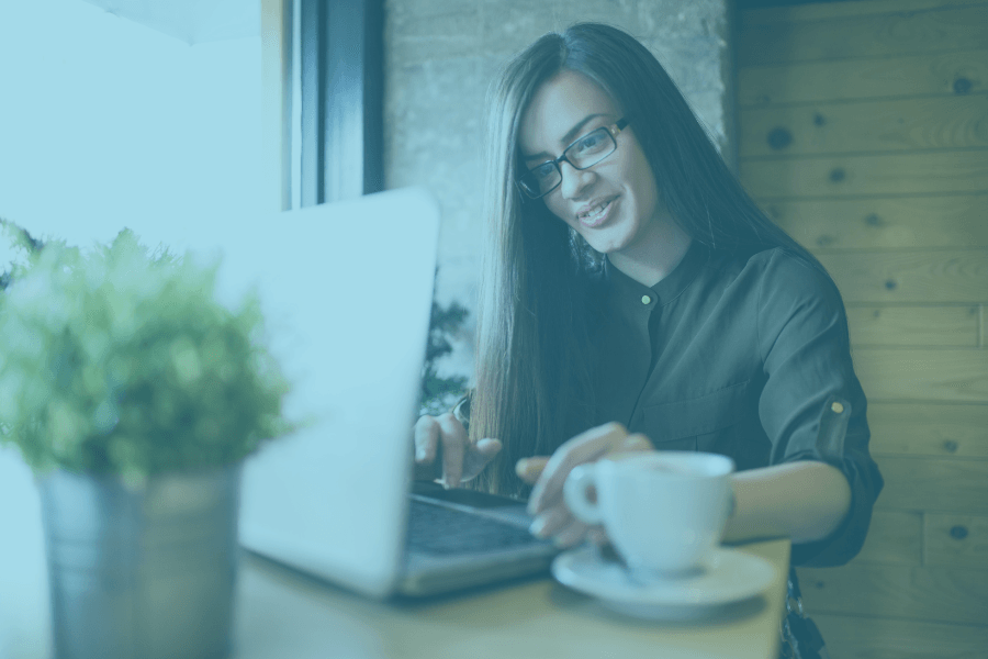 Woman smiling at a laptop with coffee in a cozy cafe, wearing glasses and a dark sweater