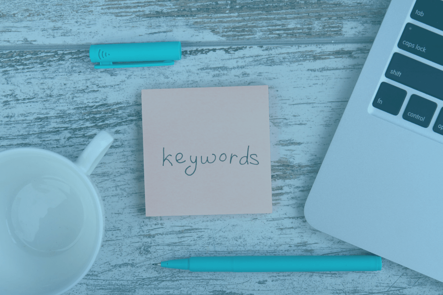 Desk with sticky note labeled “keywords,” laptop, coffee cup, and blue marker on a light wood surface