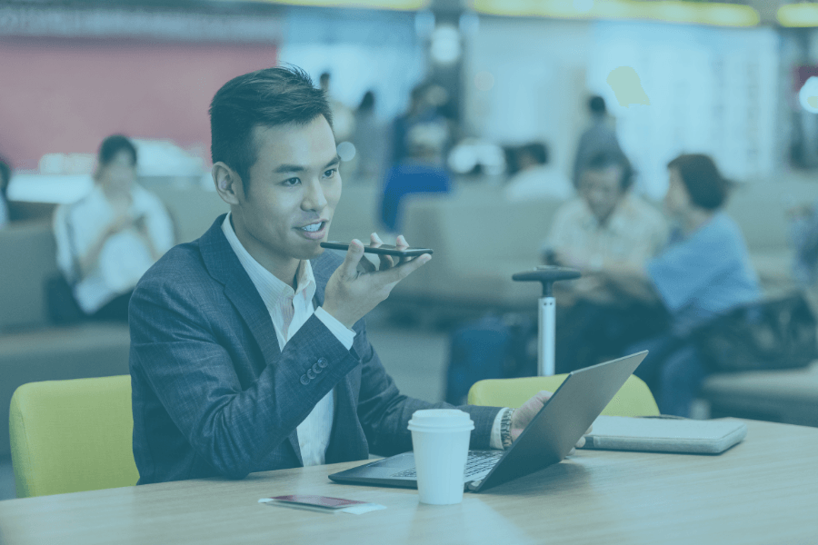 Man speaking into a phone at a desk in a busy office lounge, with a laptop and coffee cup nearby