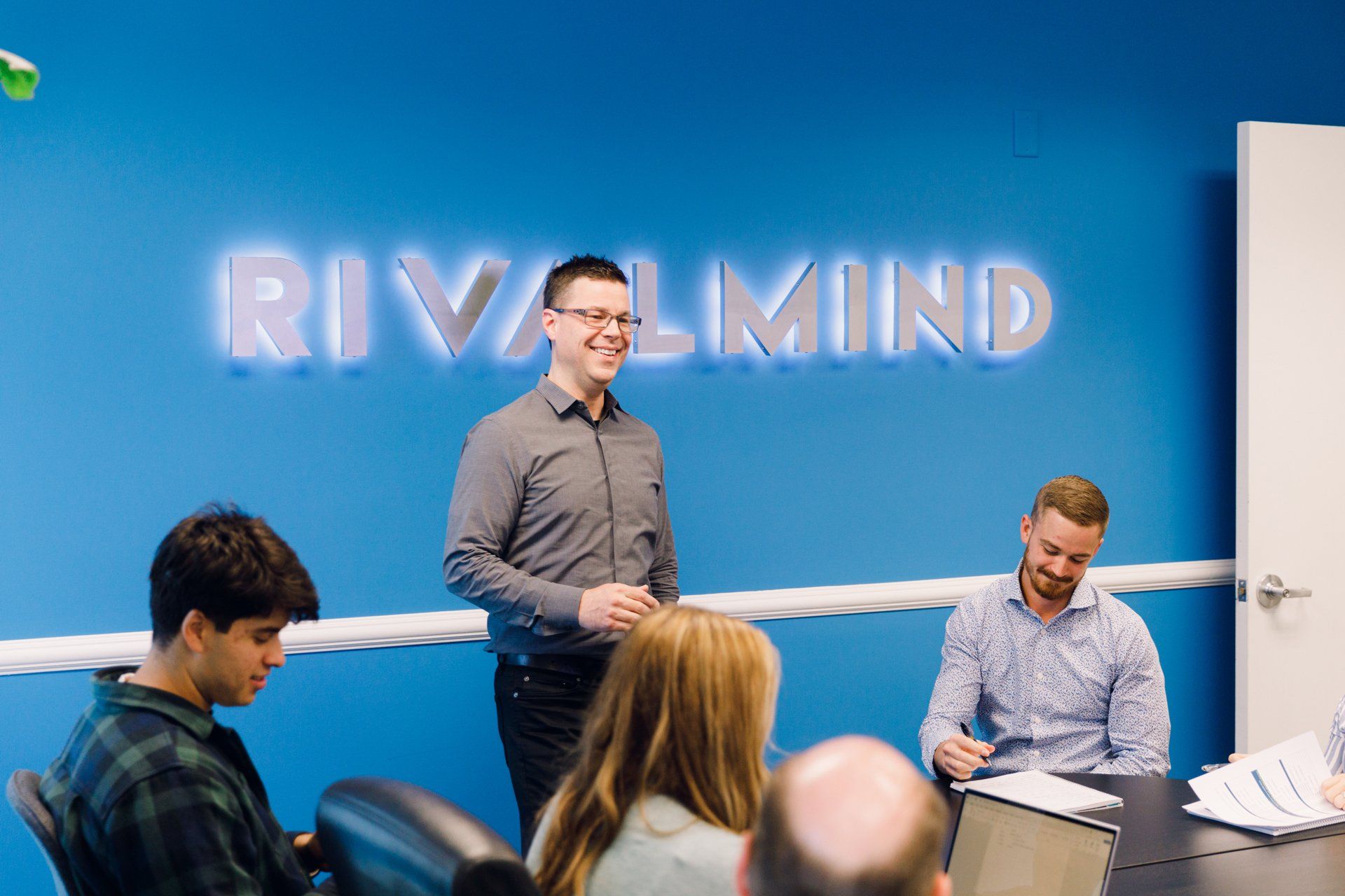 Team meeting in a blue office with a “RIVMIND” sign on the wall.