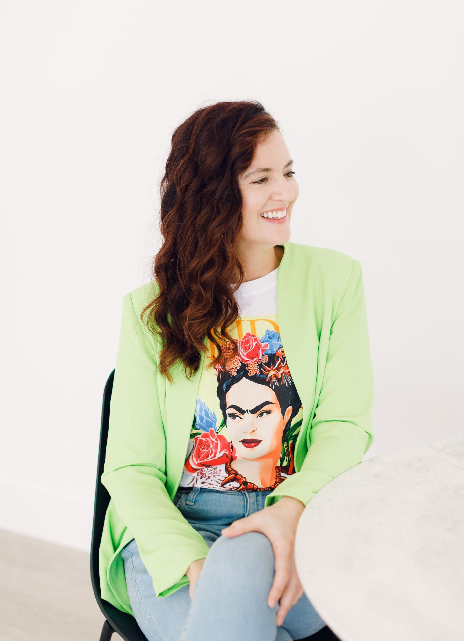 Woman sitting on a chair, smiling sideways in a lime blazer and colorful graphic T-shirt