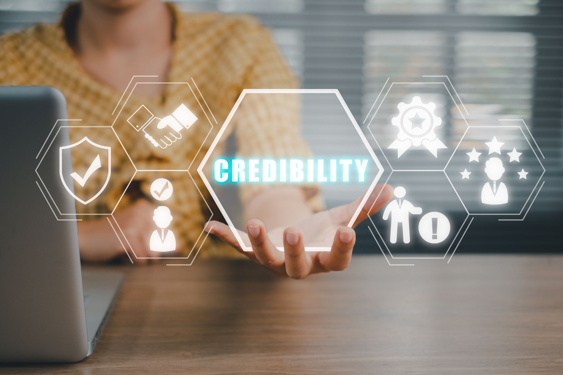Hand holding a glowing “CREDIBILITY” icon with hexagon symbols, beside a laptop on a wooden desk