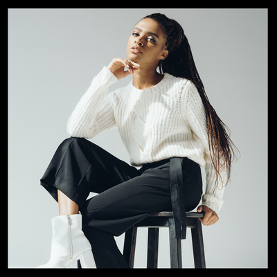 Woman seated on a stool in a white sweater and black pants, posing against a light gray background