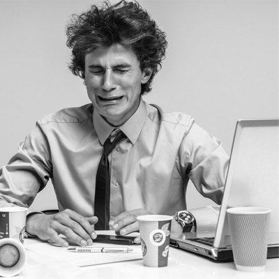 Person at desk grimacing with eyes closed, using a laptop beside a coffee cup and papers.