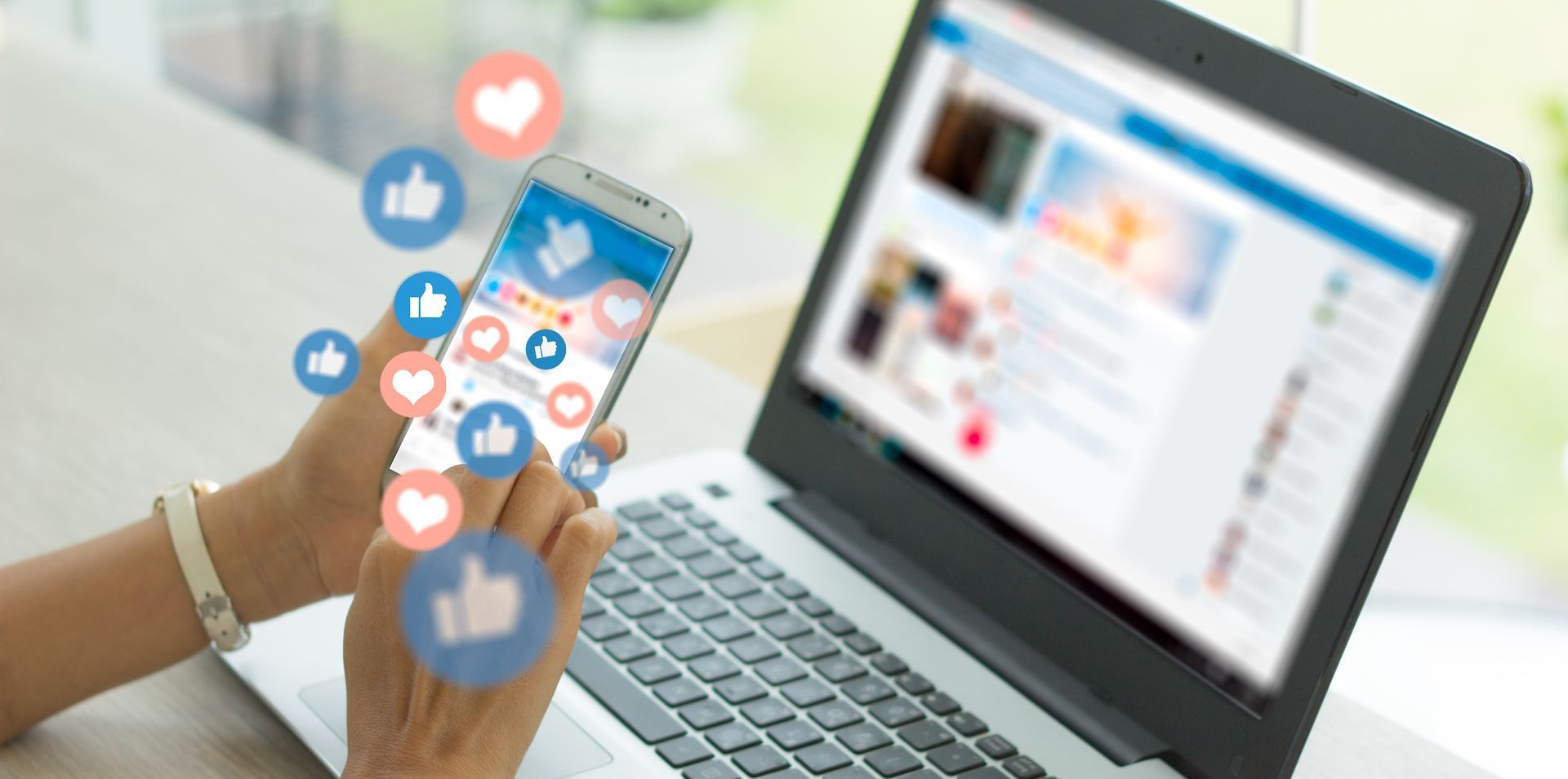 Hand holding smartphone with social media icons beside a laptop on a desk