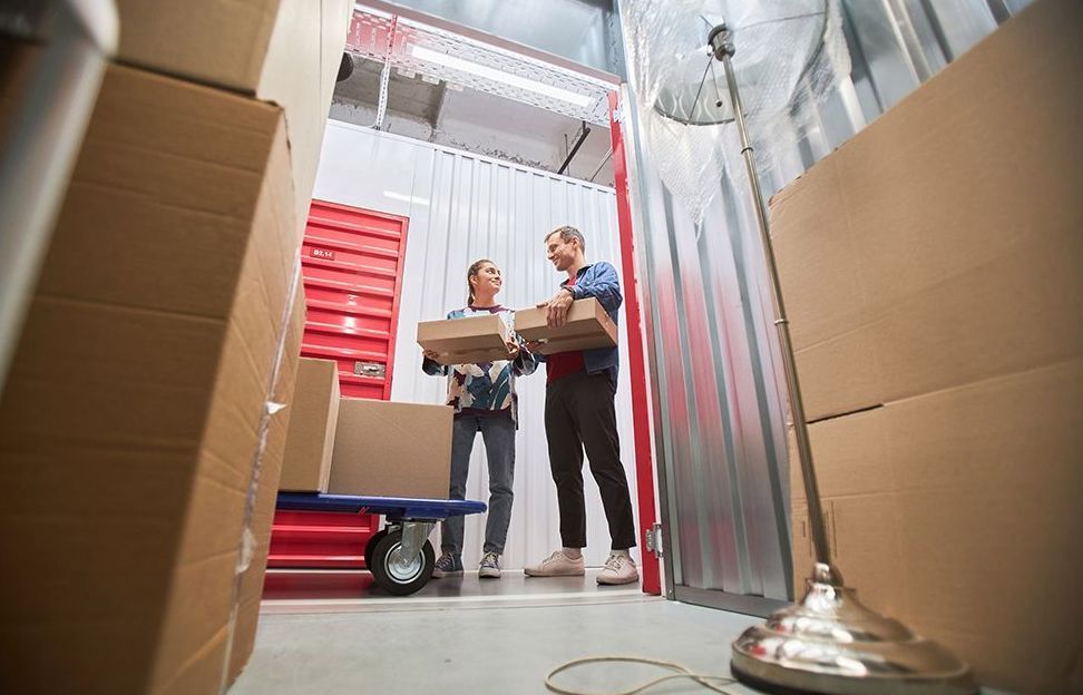 Two movers carrying a box inside a storage unit with red bins and metal walls