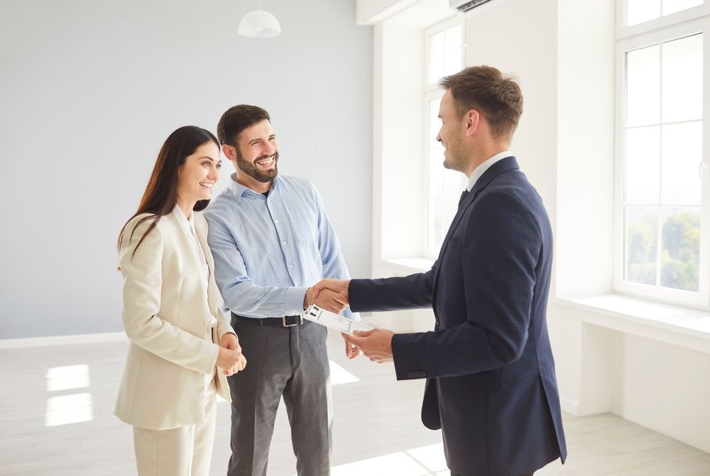 Three people meeting in a bright office, with two shaking hands while discussing a property.