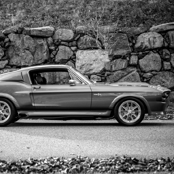 Classic sports car parked beside a stone wall on a roadside, shown in black and white.