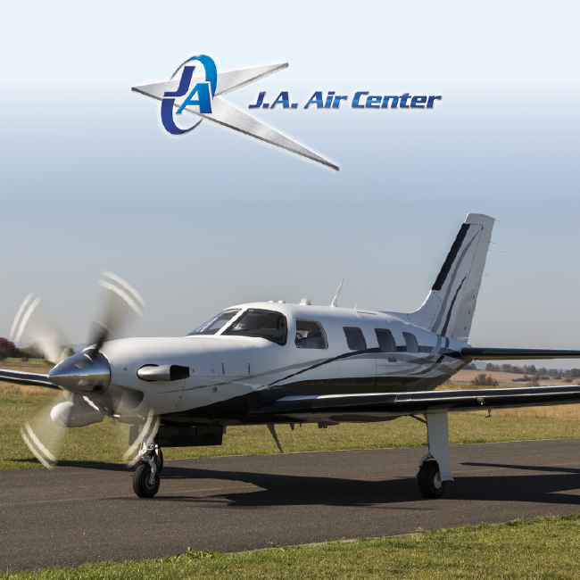 Small white propeller airplane on a runway with a J.A. Air Center logo in the sky above.