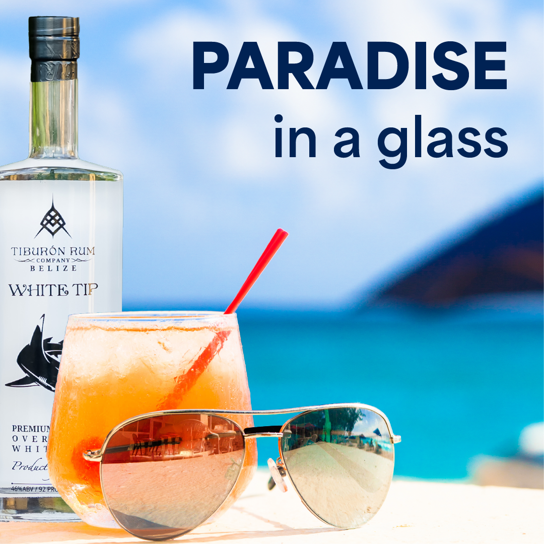 Cocktail glass and sunglasses on a beach with a white bottle and the words “Paradise in a glass.”