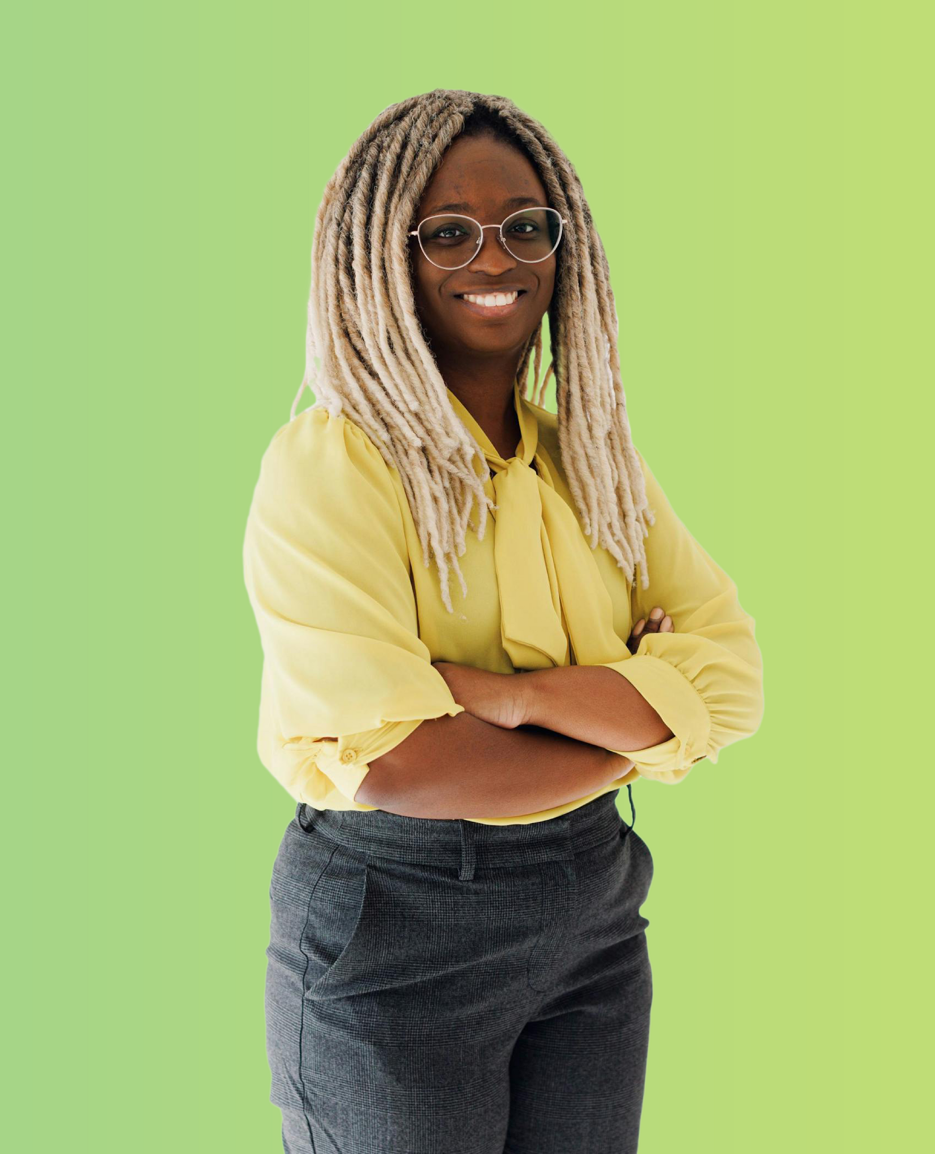 Person with glasses and blonde braids, arms crossed, wearing a yellow shirt and dark jeans against a green background