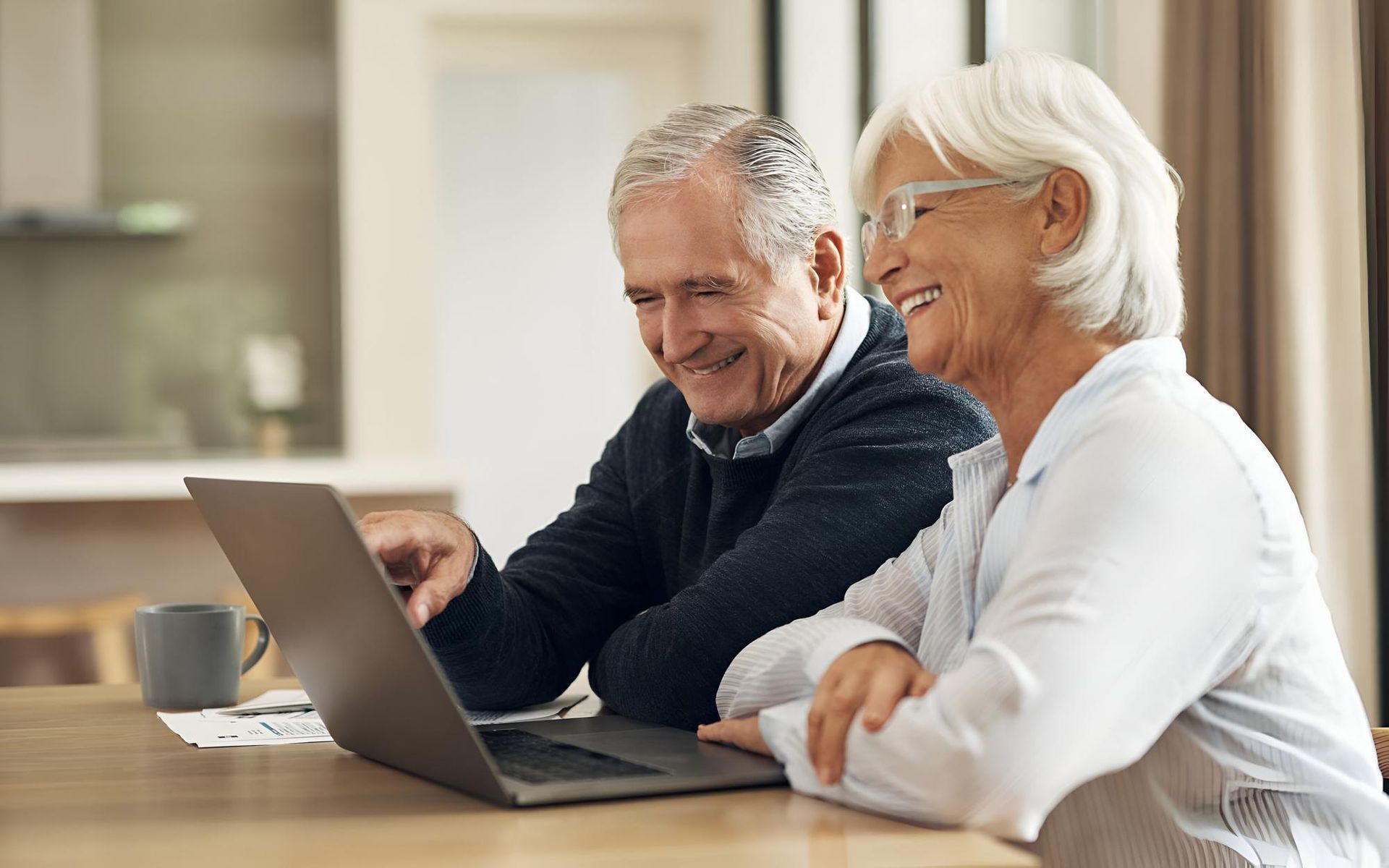 Older couple smiles while looking at a laptop computer together at a table.