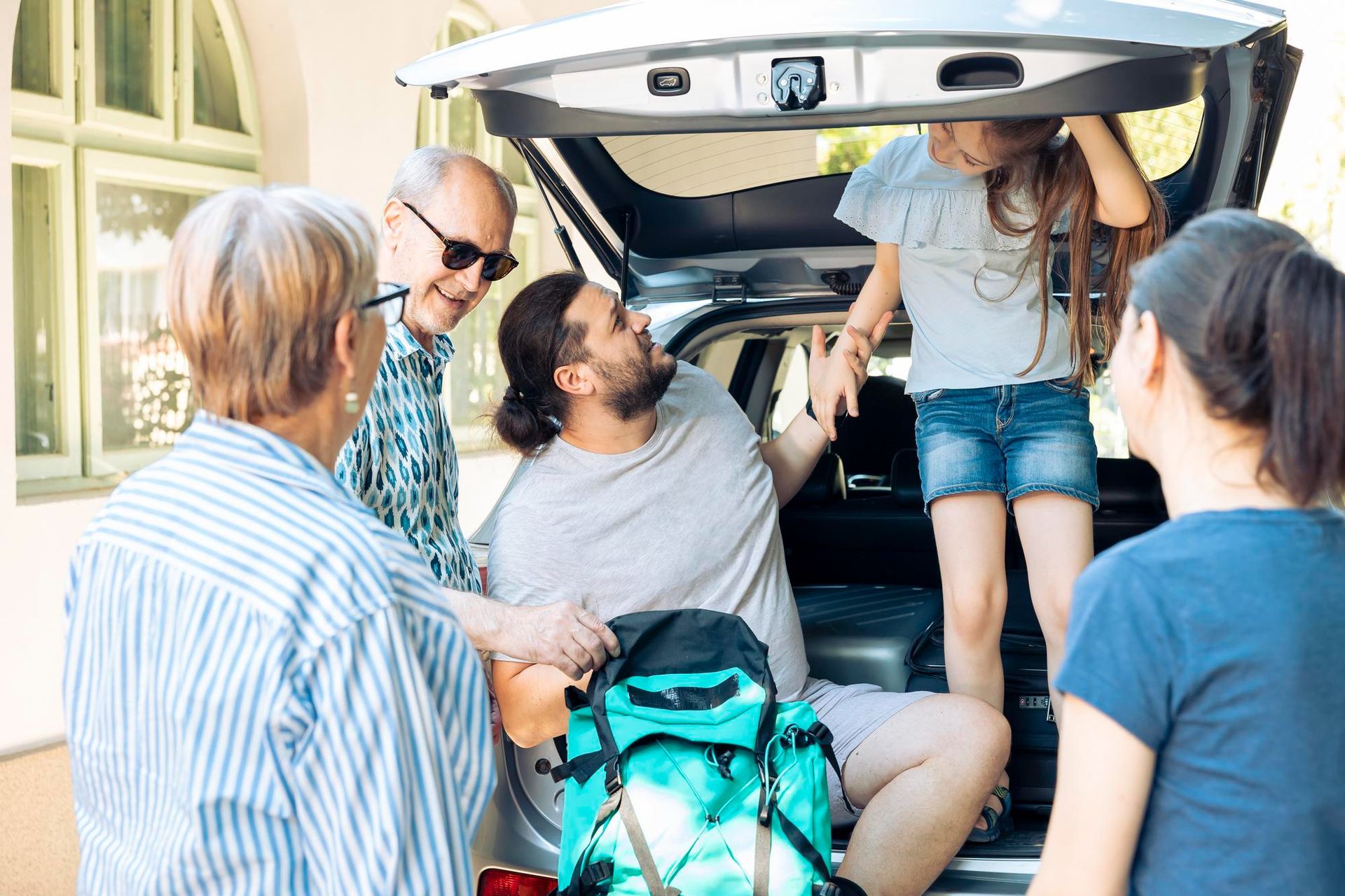 Family packing a car trunk; child and man inside, others assisting outside; sunny day.