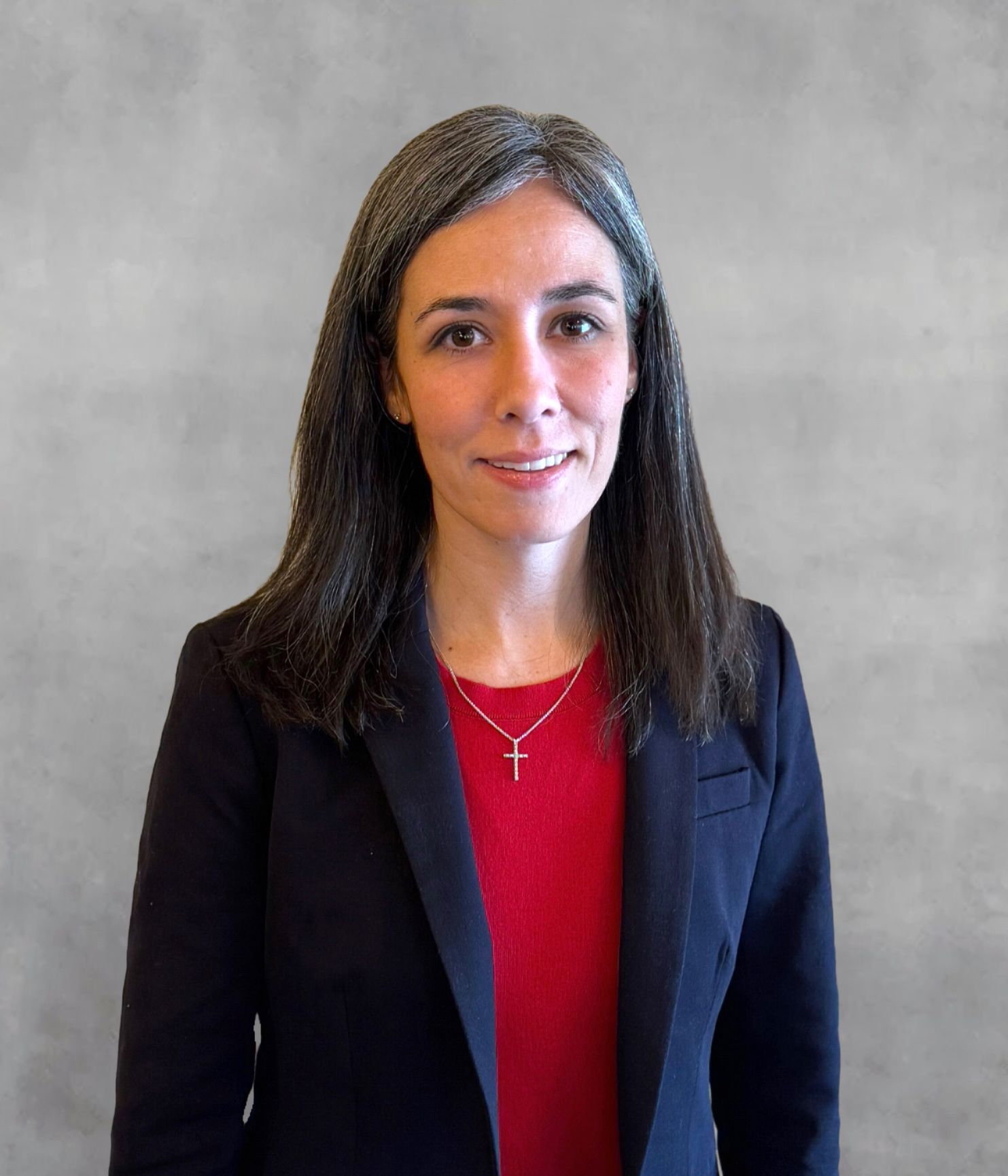Woman in dark blazer and red shirt, smiling, wearing a cross necklace. Gray background.