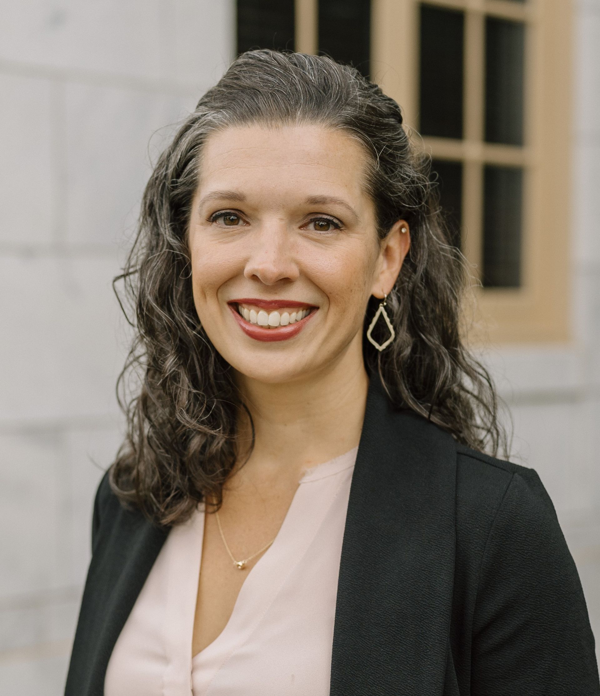 Woman with curly dark hair, smiling, wearing a black blazer and diamond-shaped earrings, outdoors.