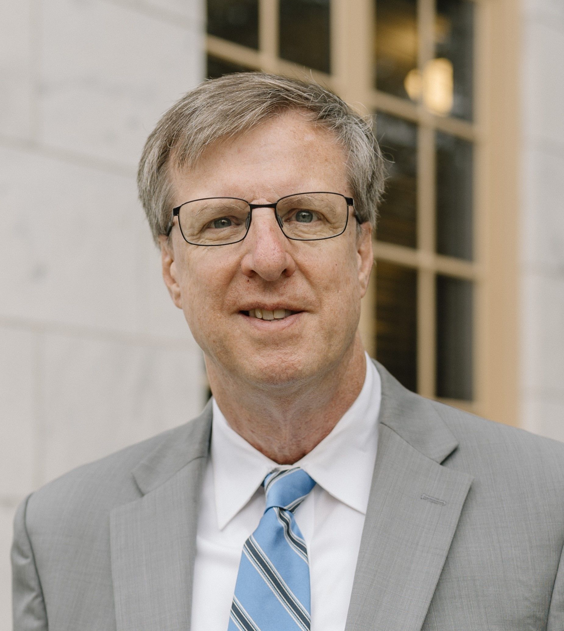 Man in glasses and suit smiling, standing in front of a light-colored building.